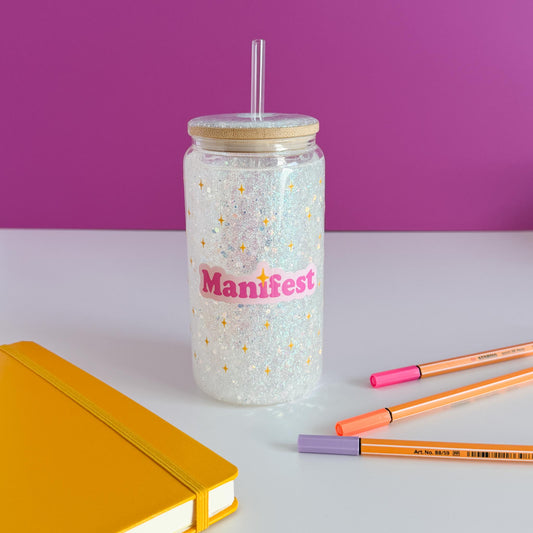 A clear glitter tumbler with a light wood lid and clear straw, featuring the word 'Manifest' in pink script with gold stars. The tumbler sits on a white surface with a yellow notebook, orange and pink pens, and a purple background.