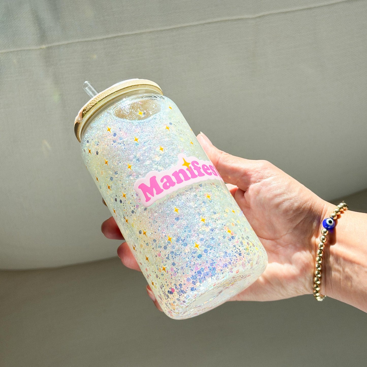 A hand holding a clear glass snow globe tumbler with a glitter lid and straw, filled with iridescent glitter and featuring a pink 'Manifest' sticker, set against a neutral gray fabric background.