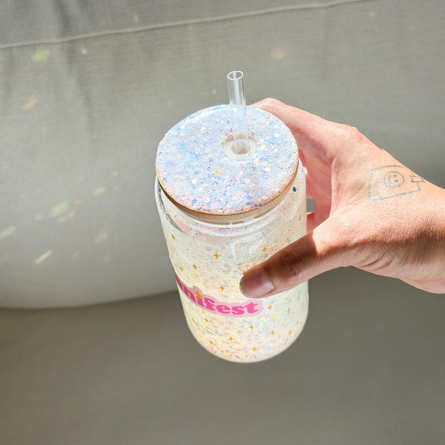 A hand holding a clear plastic tumbler with a glittery lid and straw. The tumbler has pink text reading 'manifest' and is filled with a sparkly white liquid.