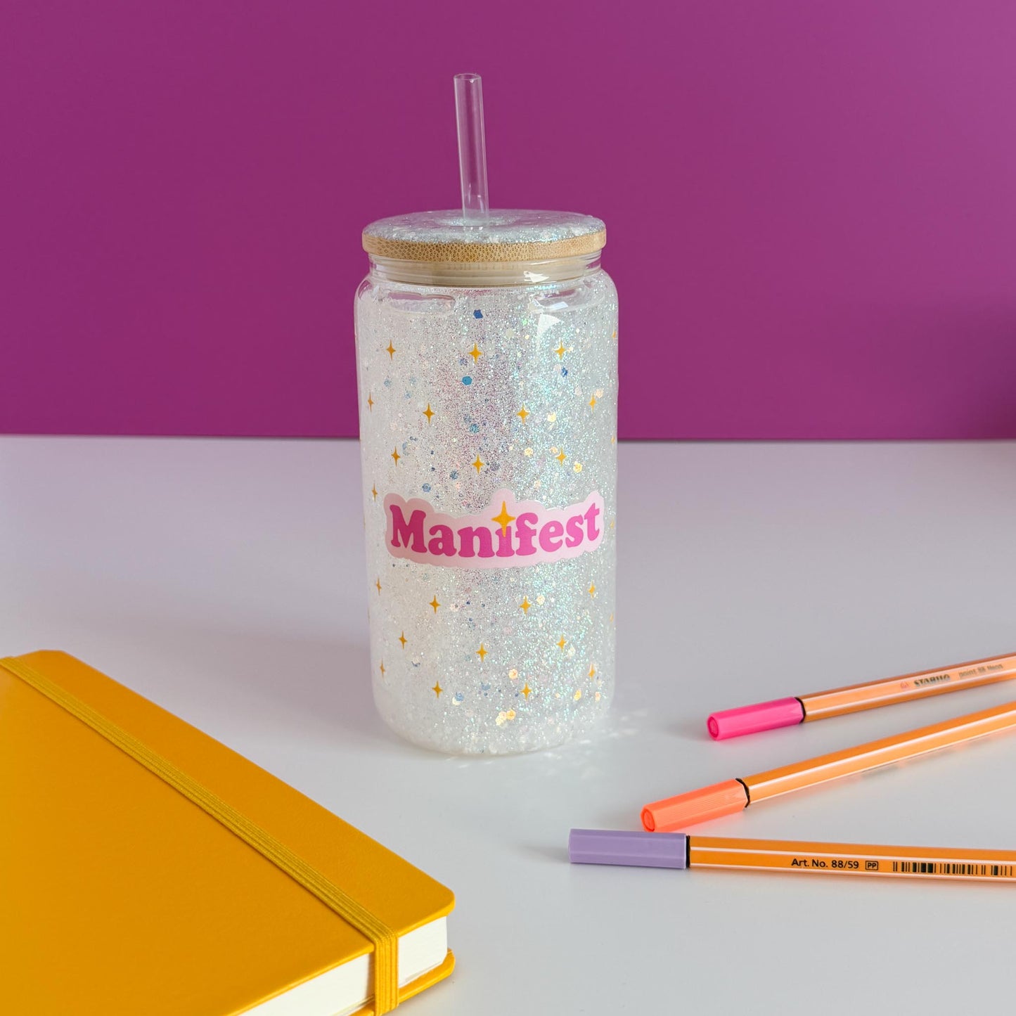 A clear glitter tumbler with a gold lid and straw, featuring the word 'Manifest' in pink on the front. The tumbler is styled on a white surface with a yellow notebook, orange and pink pens, and a purple background.