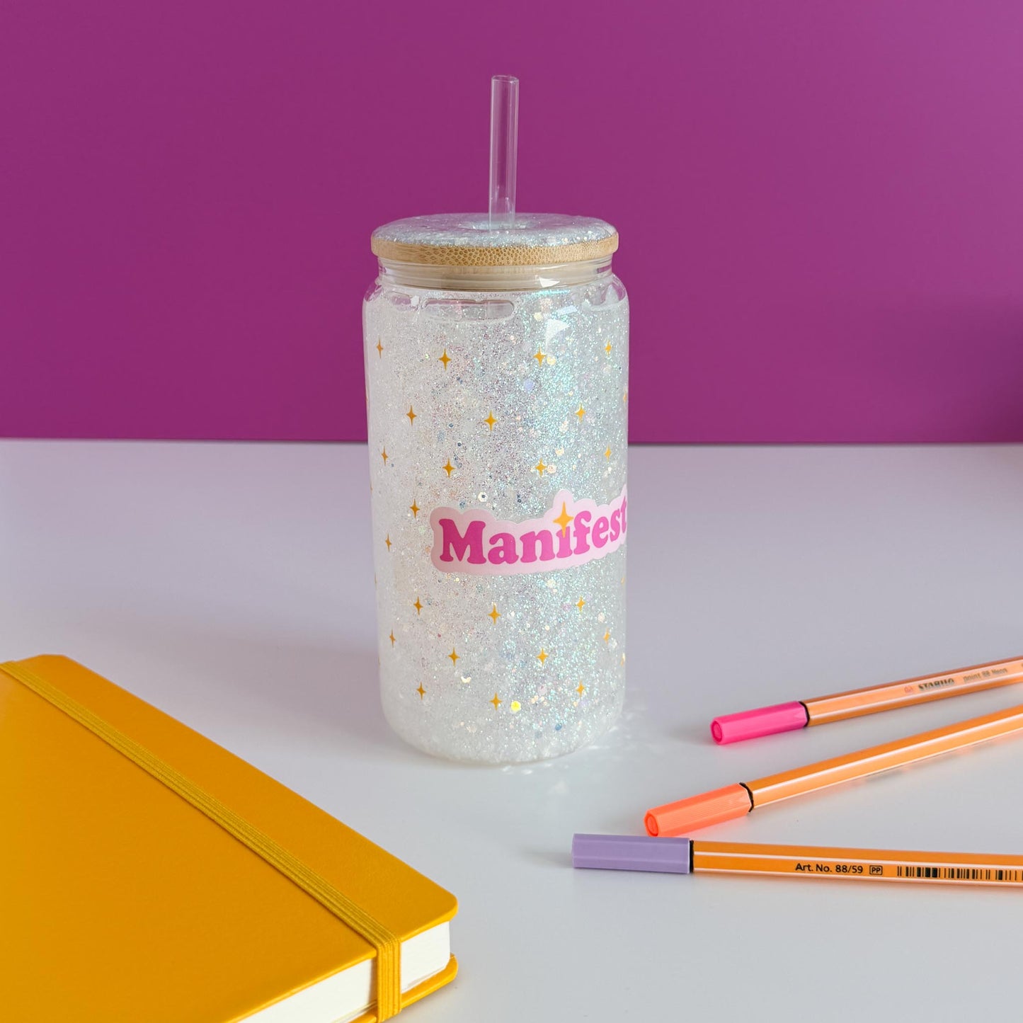 A clear glitter tumbler with a bamboo lid and straw, featuring the word 'Manifest' in pink with gold stars. The tumbler is styled on a white surface with a yellow notebook, orange and pink pens, against a purple background.