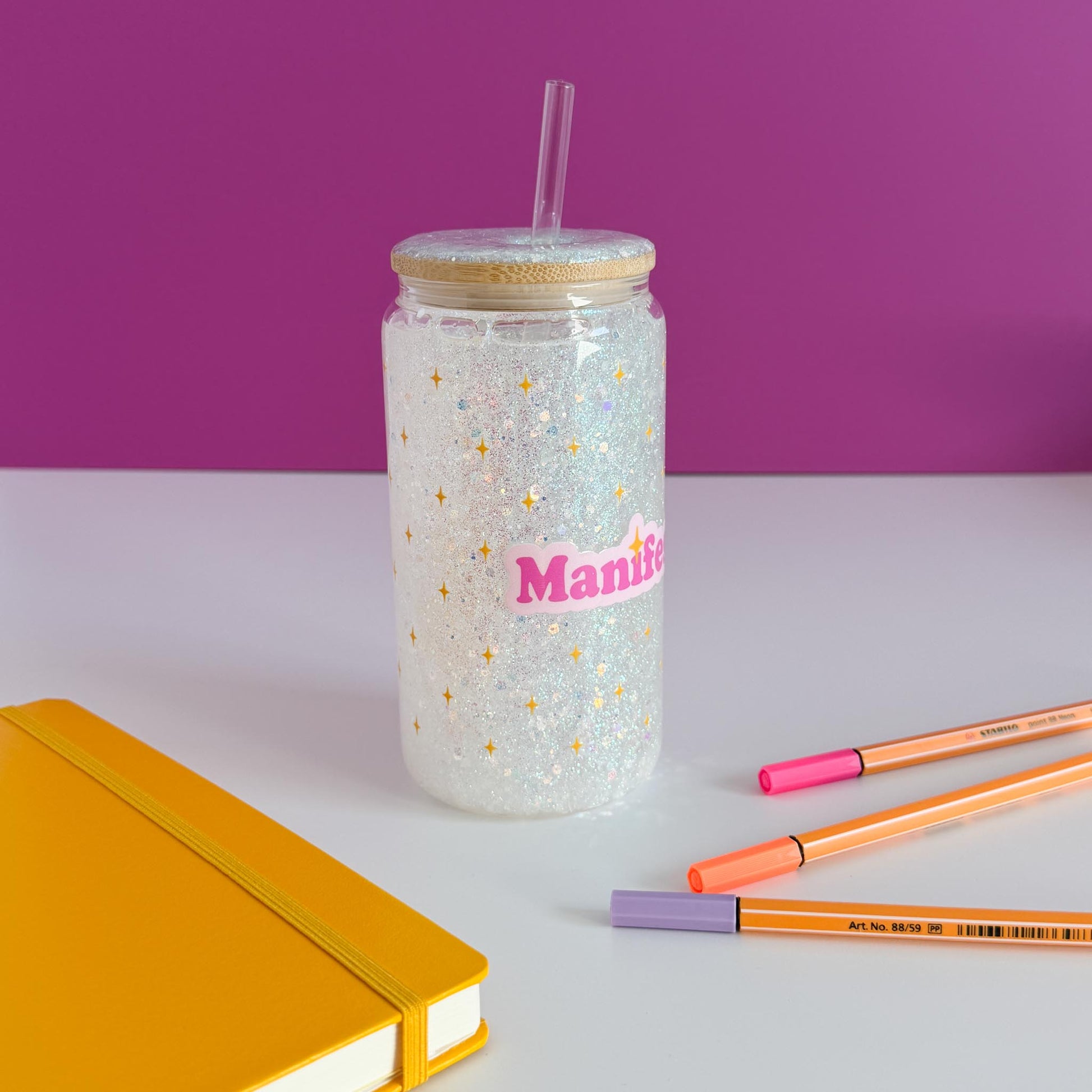 A clear glitter tumbler with a bamboo lid and straw, featuring the word 'Manifest' in pink with gold stars. The tumbler is styled on a white surface with a yellow notebook, orange and pink pens, against a purple background, rotated counter-clockwise.