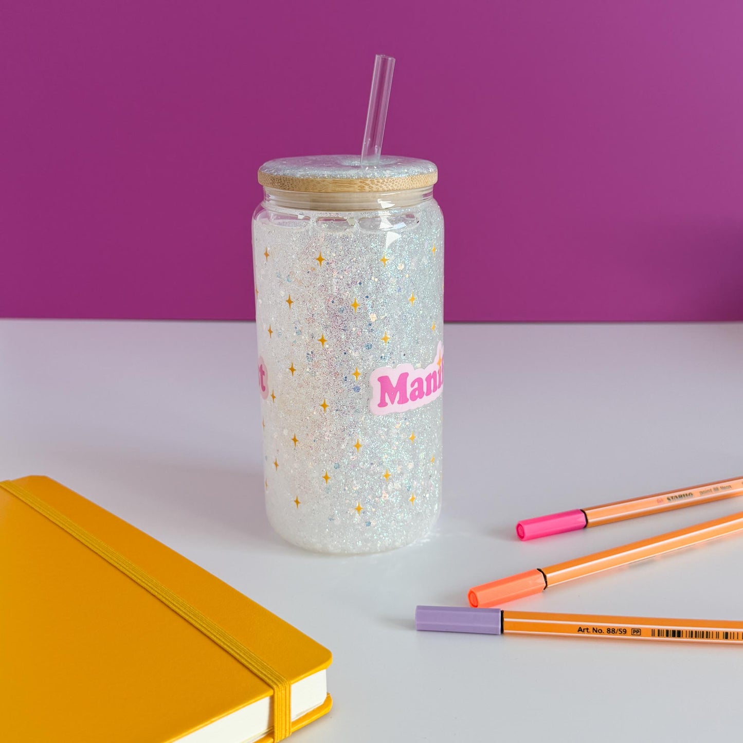 A clear glitter tumbler with a bamboo lid and straw, featuring the word 'Manifest' in pink with gold stars. The tumbler is styled on a white surface with a yellow notebook, orange and pink pens, against a purple background, rotated counter-clockwise.