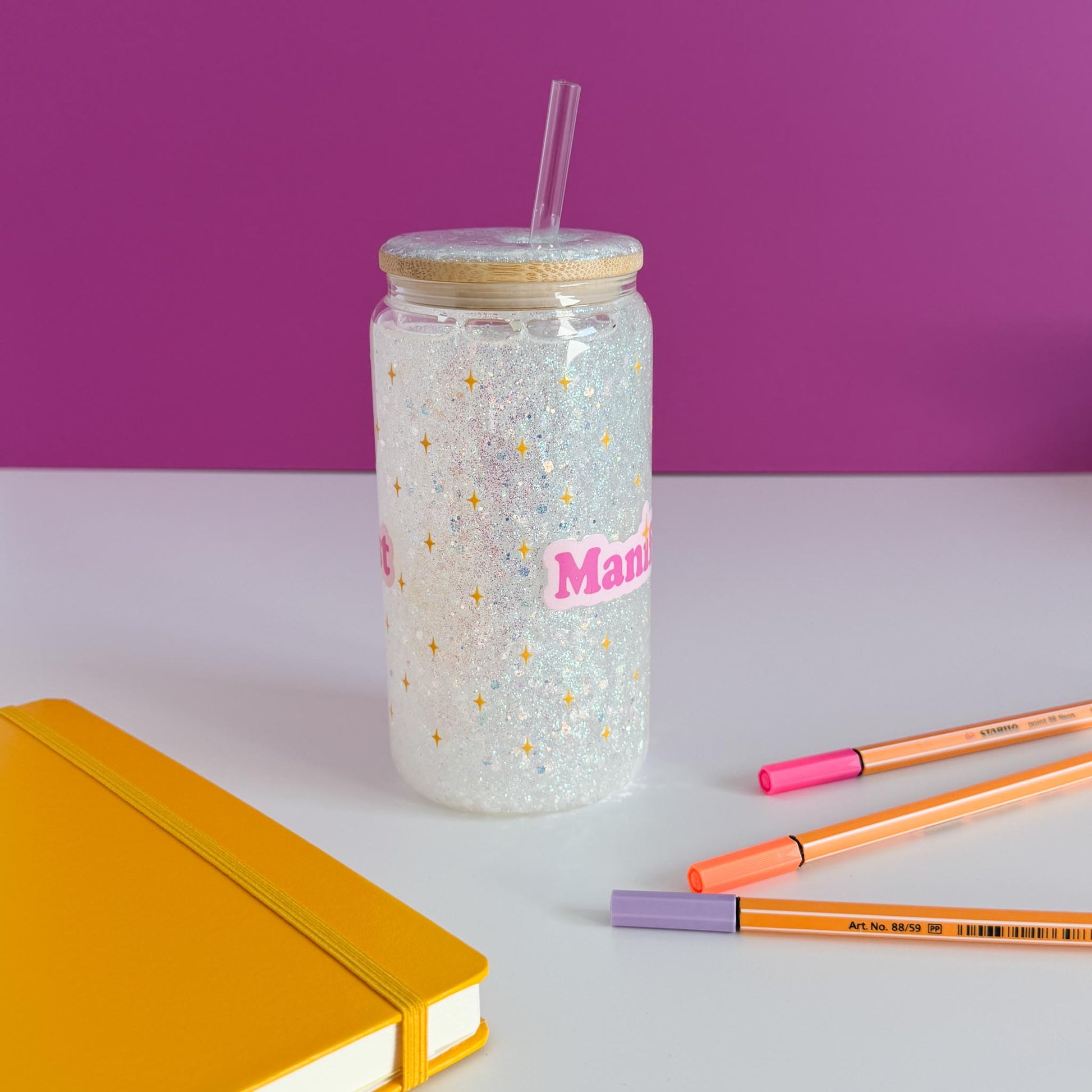 A clear glitter tumbler with a bamboo lid and straw, featuring the word 'Manifest' in pink with gold stars. The tumbler is styled on a white surface with a yellow notebook, orange and pink pens, against a purple background, rotated counter-clockwise.