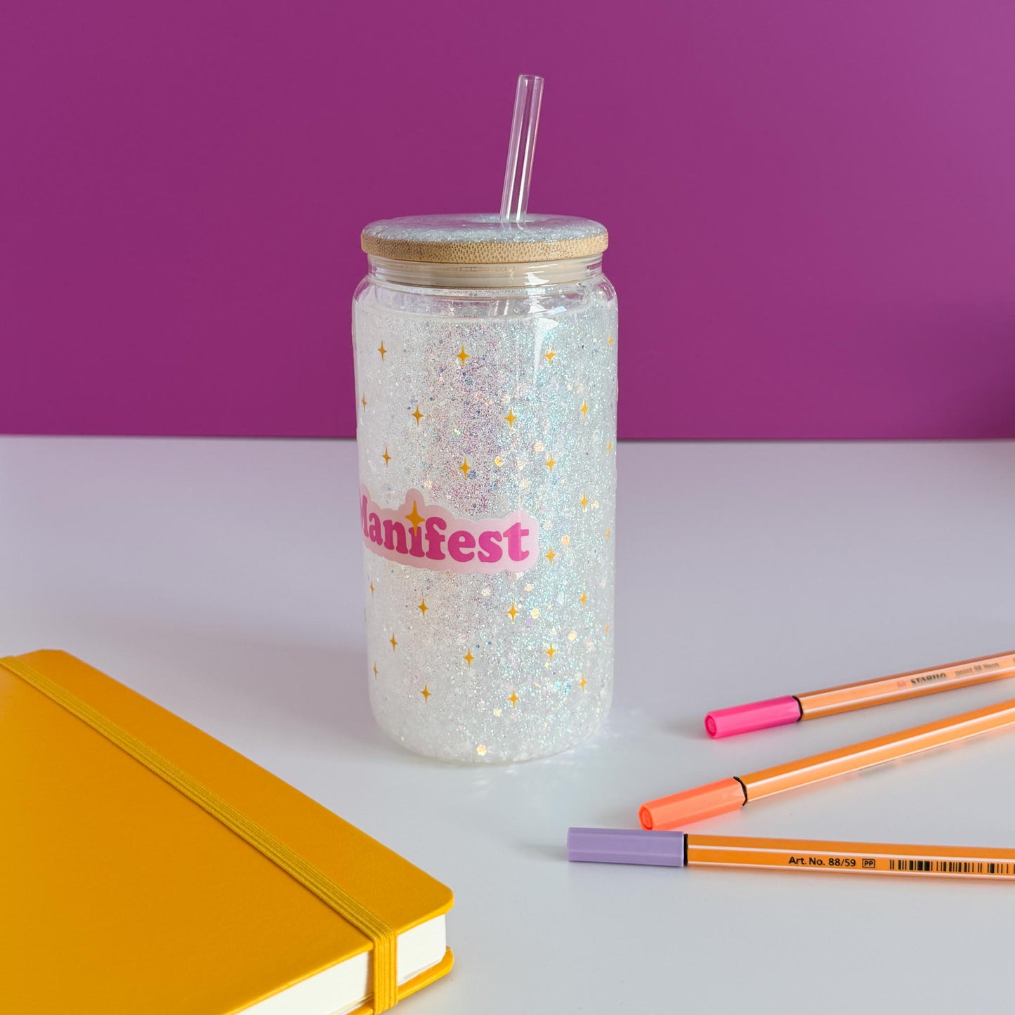 A clear glitter tumbler with a bamboo lid and straw, featuring the word 'Manifest' in pink with gold stars. The tumbler is styled on a white surface with a yellow notebook, orange and pink pens, against a purple background, rotated counter-clockwise.