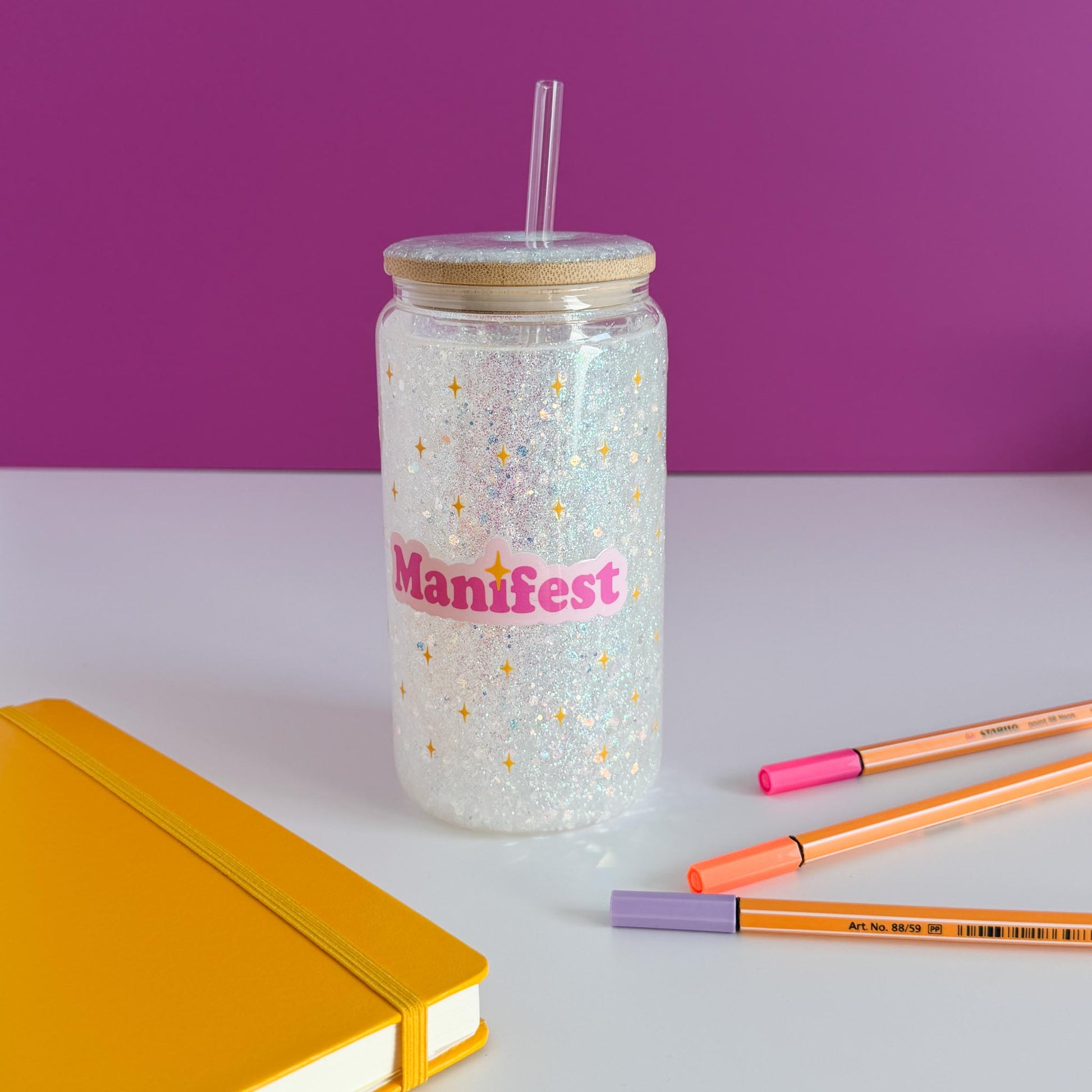 A clear glitter tumbler with a bamboo lid and straw, featuring the word 'Manifest' in pink with gold stars. The tumbler is styled on a white surface with a yellow notebook, orange and pink pens, against a purple background, rotated counter-clockwise.