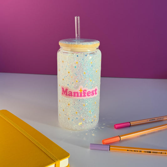 A clear glitter tumbler with a bamboo lid and straw, featuring a pink 'Manifest' sticker, styled on a white surface with a purple background.