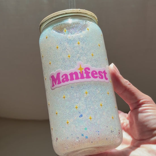 A video of a hand holding a clear glass snow globe tumbler with a glitter lid and straw, filled with iridescent glitter and featuring a pink 'Manifest' sticker, set against a neutral gray fabric background.