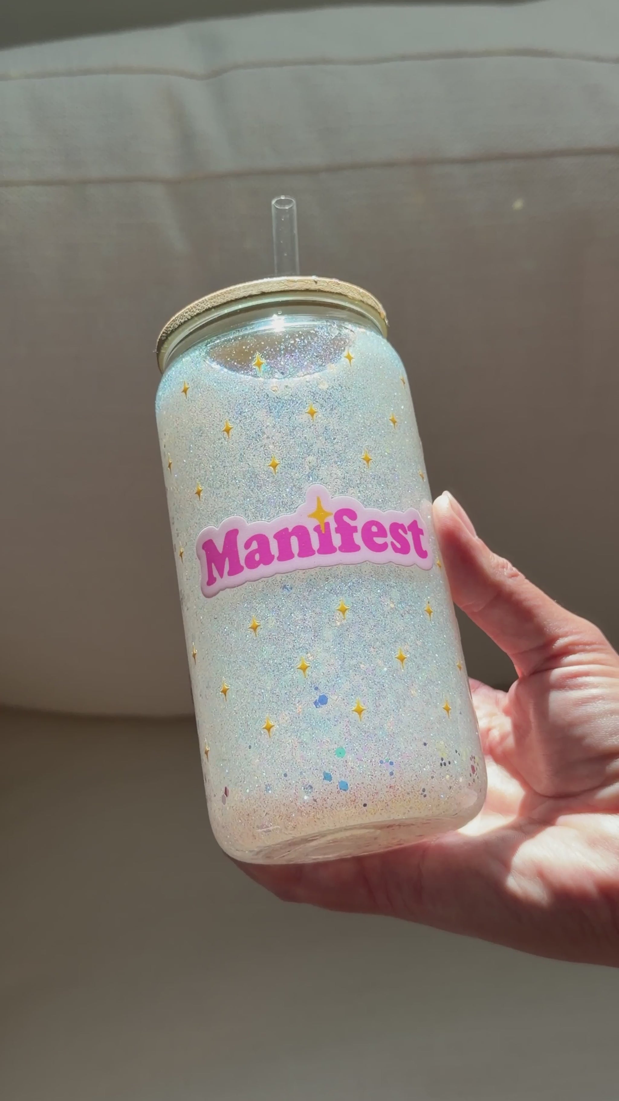 A video of a hand holding a clear glass snow globe tumbler with a glitter lid and straw, filled with iridescent glitter and featuring a pink 'Manifest' sticker, set against a neutral gray fabric background.